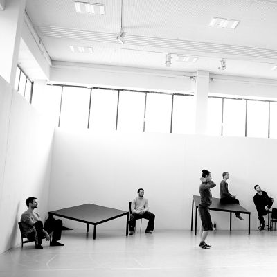 The space is large, light, and minimalist, with white walls, a high, white ceiling. Several individuals are scattered in the space. They appear to be involved in a dance or movement performance. Some are standing, while others are seated on or near tables. The individuals are dressed in dark clothing. There are a few simple, rectangular black metal tables of varying heights within the space. The tables serve as a backdrop and props for the performance.
