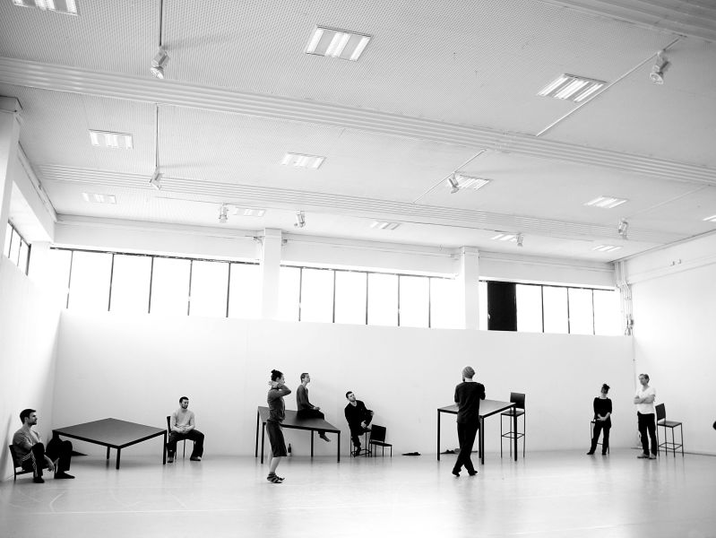 The space is large, light, and minimalist, with white walls, a high, white ceiling. Several individuals are scattered in the space. They appear to be involved in a dance or movement performance. Some are standing, while others are seated on or near tables. The individuals are dressed in dark clothing. There are a few simple, rectangular black metal tables of varying heights within the space. The tables serve as a backdrop and props for the performance.